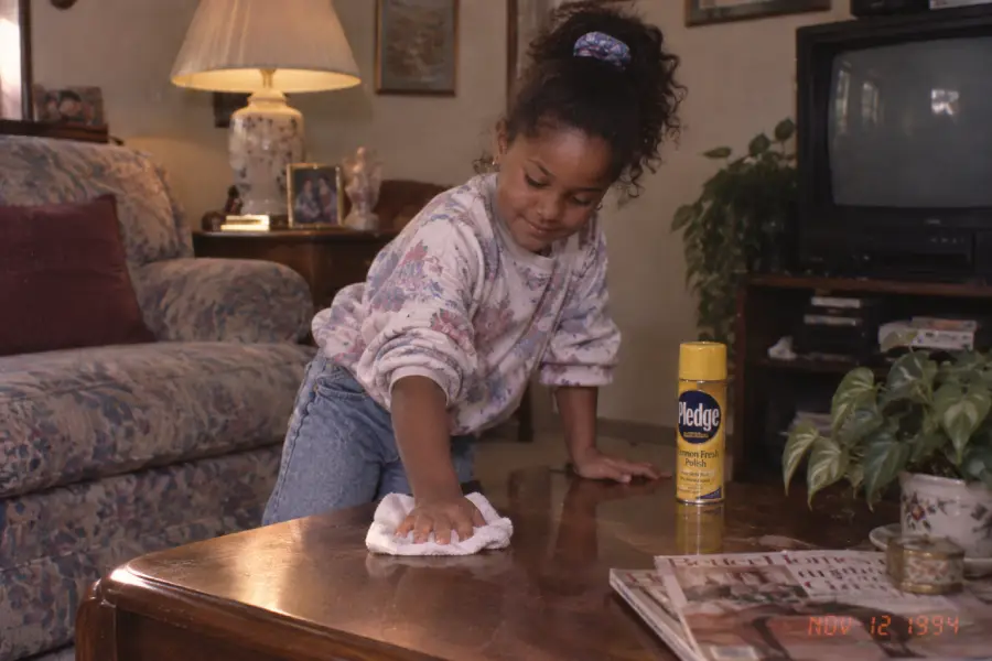 A young girl cleans a wooden coffee table with a cloth. A can of Pledge polish sits nearby. The room has a floral couch, TV, lamp, and dated decor. The photo is timestamped November 12, 1994.