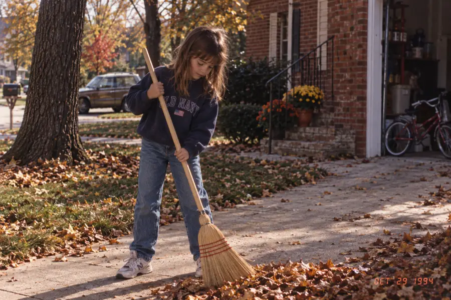 A young girl sweeps fallen autumn leaves on a driveway in front of a house. She wears jeans and a navy sweatshirt. There are colorful mums and a bicycle near the open garage. The trees have autumn-colored leaves.