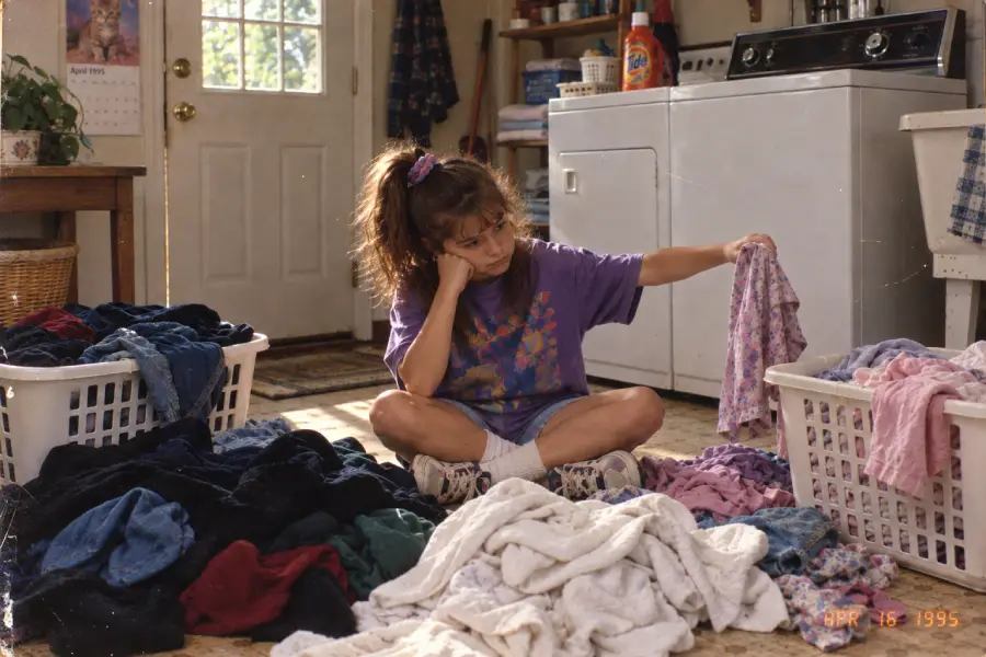 A young girl sits on the floor surrounded by laundry, looking bored as she holds up a shirt. Behind her are a washing machine, dryer, laundry detergent, and a door with sunlight coming through.