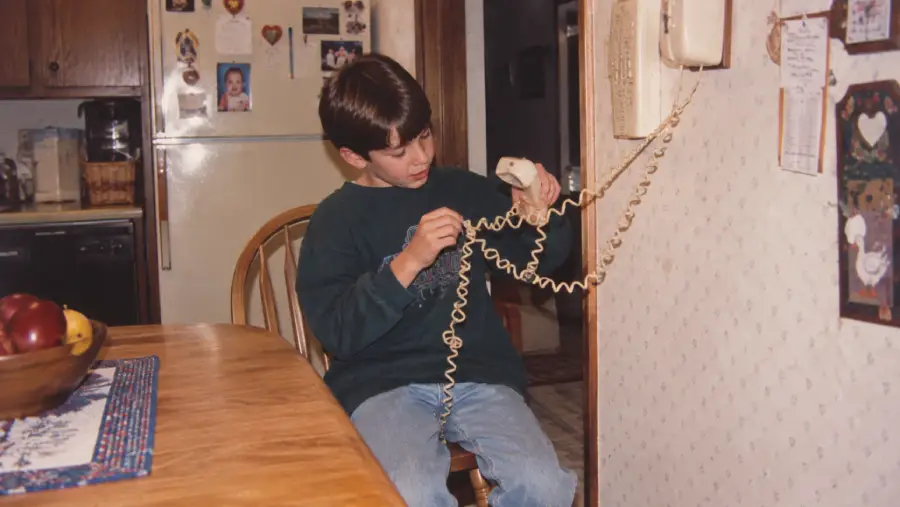 A young boy sits at a kitchen table, tangled in the cord of a wall-mounted landline phone, looking at the receiver in his hand. The kitchen has wood cabinets and a refrigerator covered with magnets and photos.