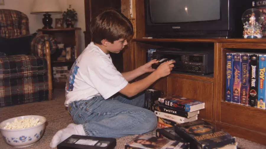 A boy kneels by a TV and VCR, inserting a VHS tape. He is surrounded by VHS tapes and a bowl of popcorn on the carpet in a cozy living room.