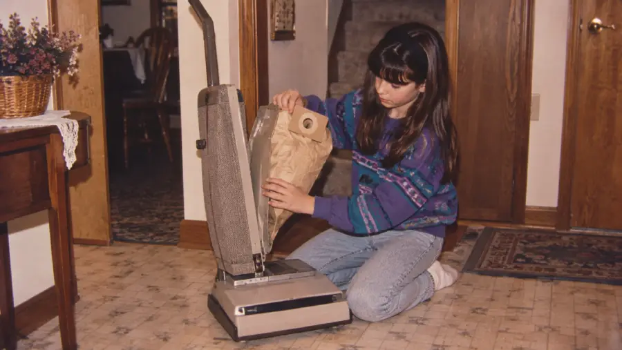 A girl kneels on a kitchen floor, replacing a vacuum cleaner bag. She wears a blue patterned sweater and jeans. The room has wooden doors, a carpet, and a table with a flower basket.