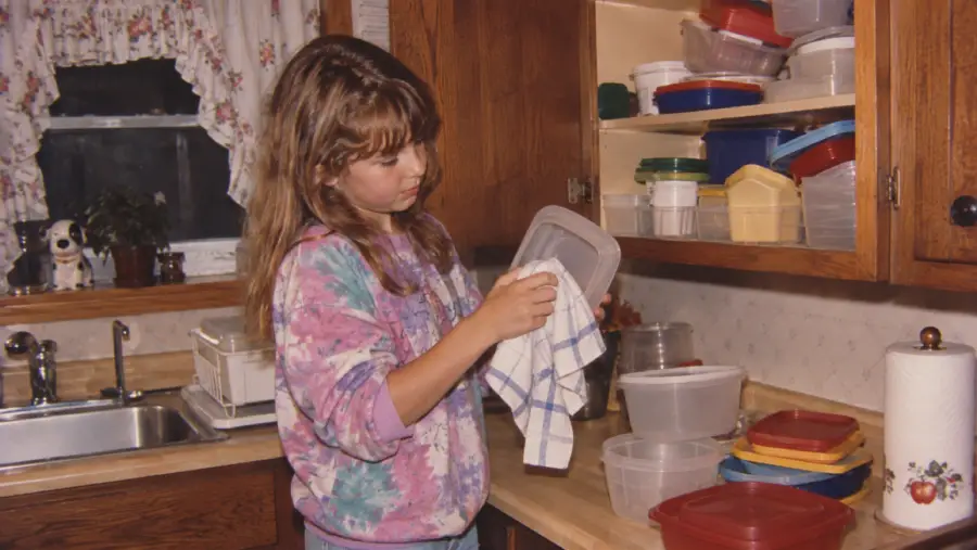 A girl in a colorful sweatshirt dries a plastic container with a towel in a kitchen, standing near a sink and open cabinet filled with various food storage containers.