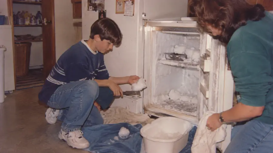 Two people are defrosting a freezer, removing chunks of ice from the shelves. A blue towel and white bin are on the floor to catch water and ice. The freezer door is open, revealing frost buildup inside.