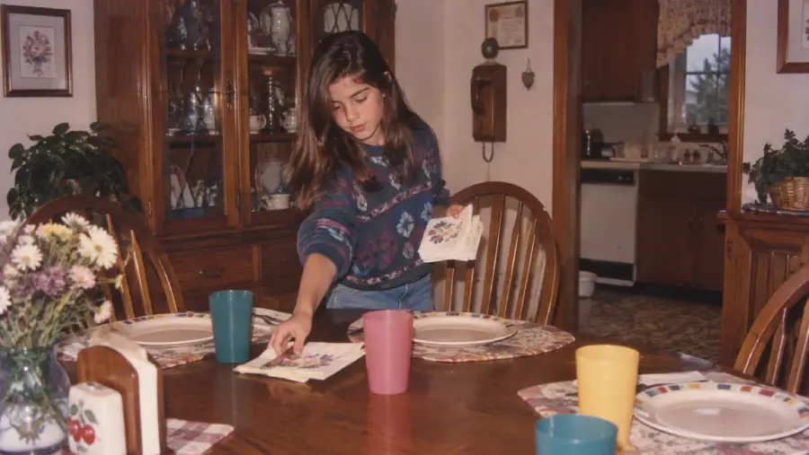 A young girl sets the dining table with plates, cups, and napkins in a cozy, wood-furnished kitchen. Flowers and placemats decorate the table.