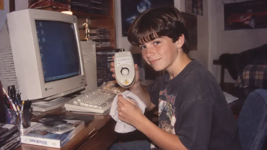 A boy sits at a desk with a vintage computer, holding and cleaning a mouse. Papers, pens, and computer accessories are nearby. He smiles at the camera in a cozy, cluttered room.