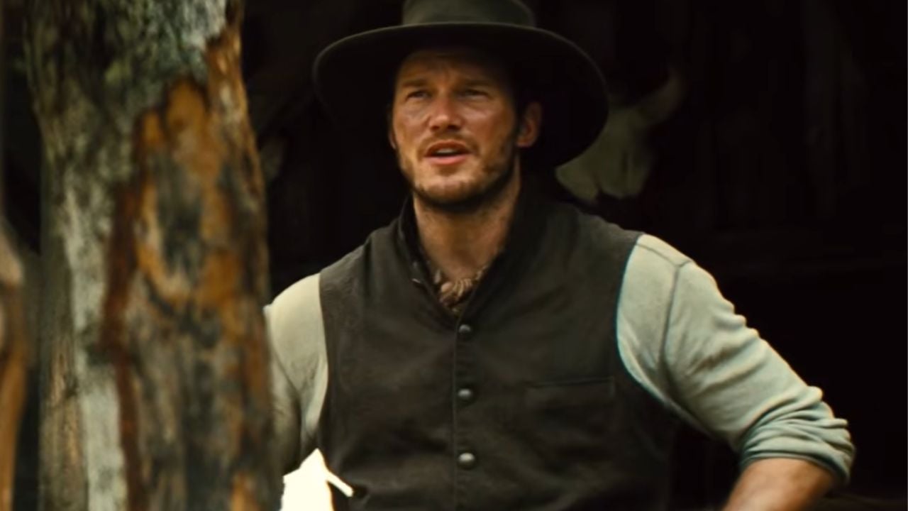 A man wearing a wide-brimmed hat and vest stands outdoors near a tree, looking into the distance with a serious expression.