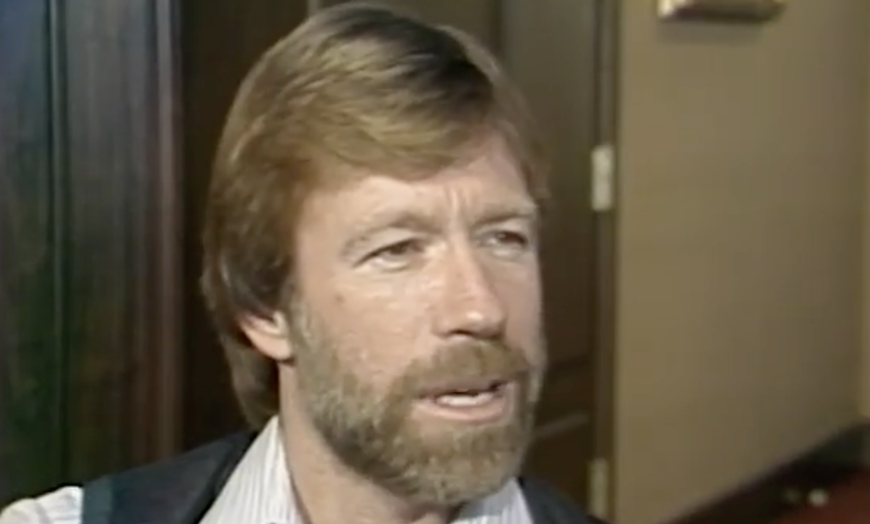 A bearded man with light brown hair and mustache is speaking indoors, wearing a striped shirt and a dark vest. The background shows a brown door and beige walls.
