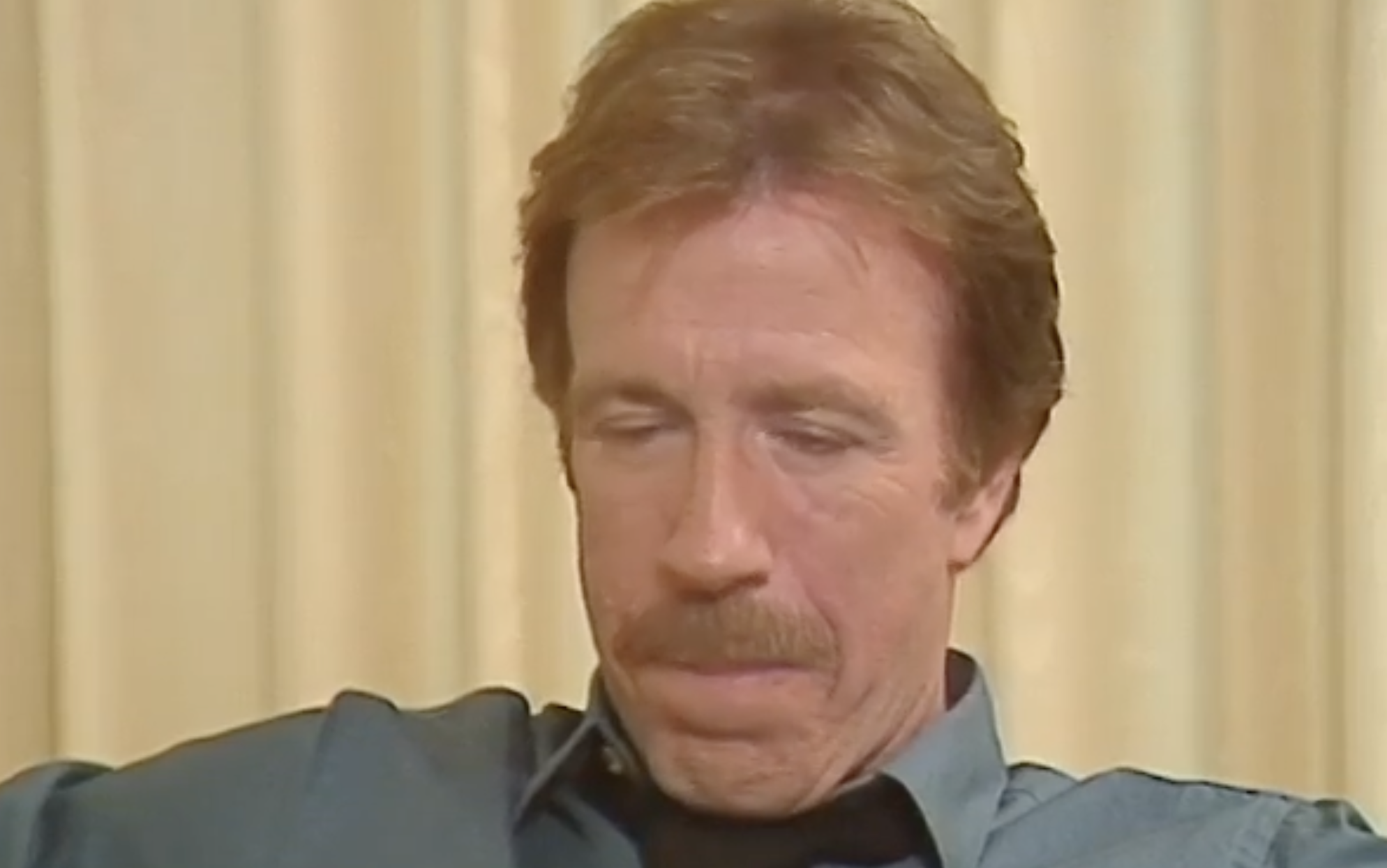 Man with light brown hair and mustache wearing a blue collared shirt looks down with a neutral expression. Cream-colored curtains are in the background.
