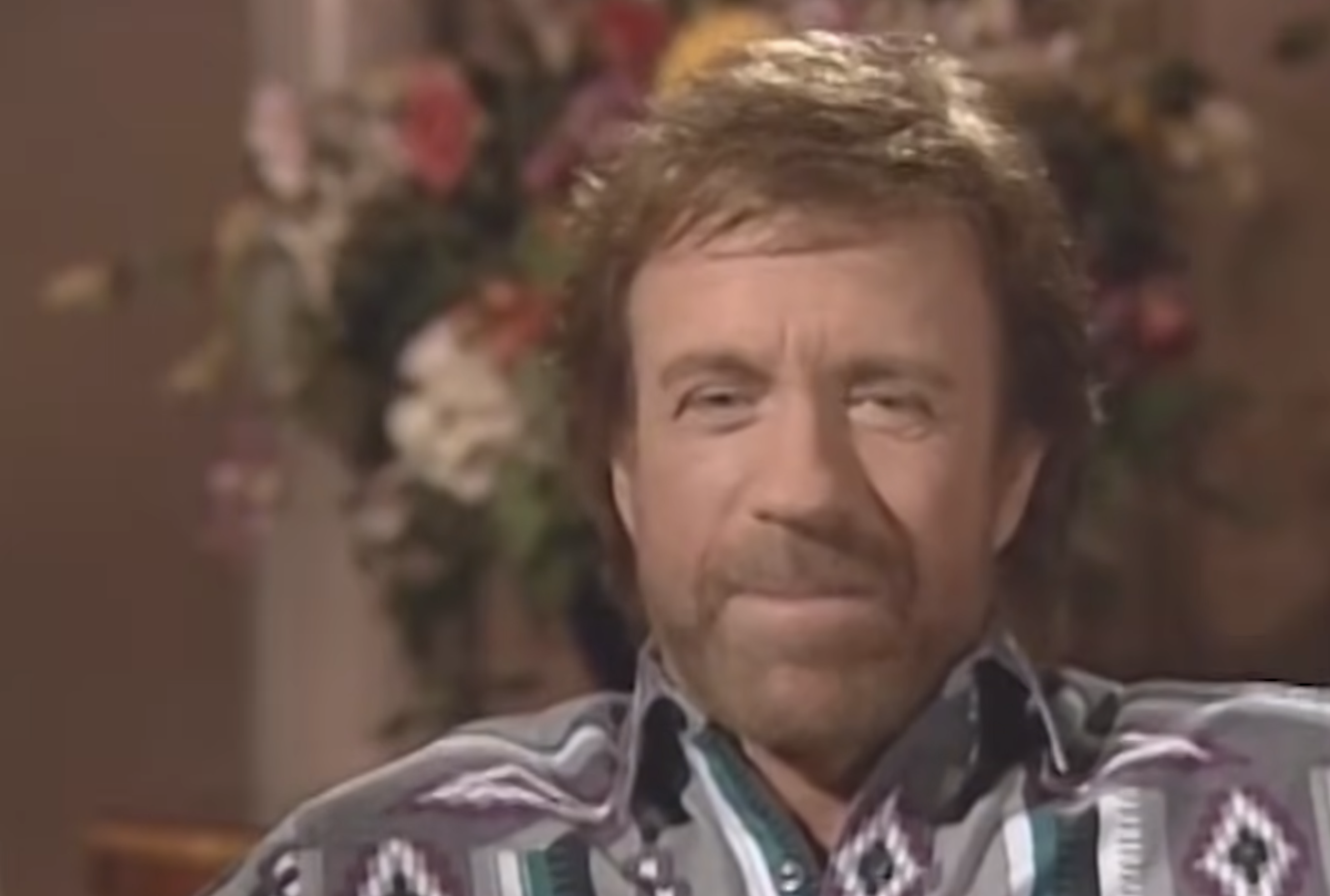 A man with a beard and mustache sits indoors, wearing a patterned shirt. There is a blurred bouquet of flowers in the background.