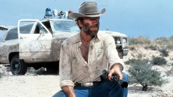 A man in a cowboy hat and badge kneels in front of a dusty police car in a desert, holding a rifle. He wears a dirty shirt and looks into the distance, with dry brush and blue sky in the background.