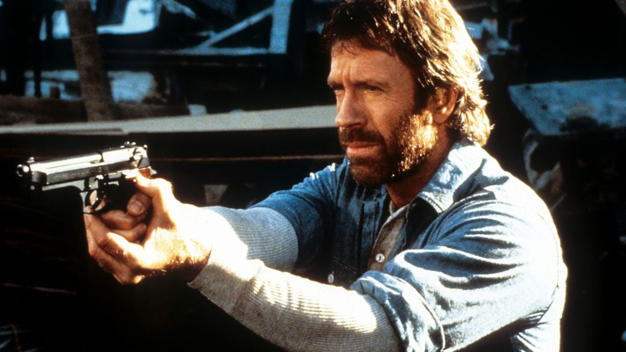 A man with a beard and mustache, wearing a denim shirt, aims a handgun with both hands while looking serious in an outdoor setting.