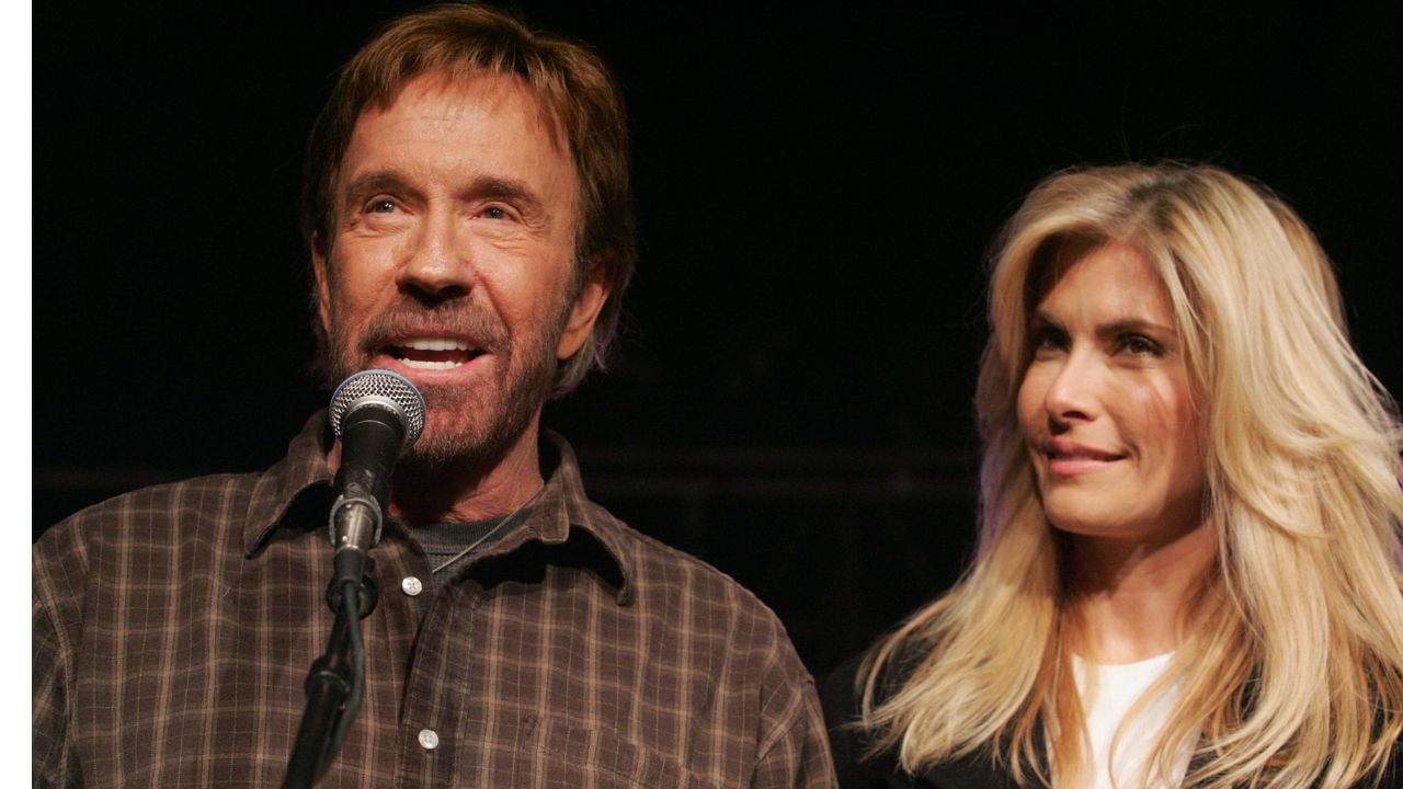 A man with a beard speaks into a microphone while a blonde woman stands beside him, both looking forward. The background is dark and the mood appears to be casual.