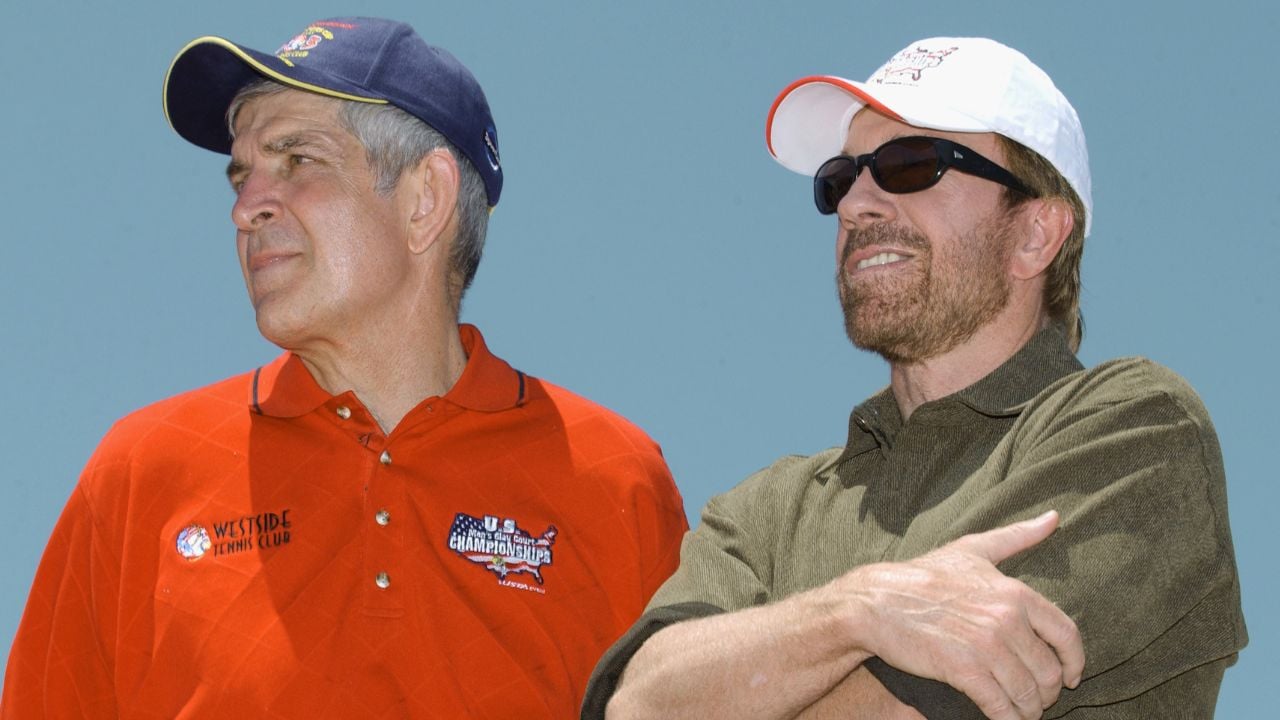 Two men stand side by side outdoors. One wears a red polo, navy cap, and looks left. The other wears sunglasses, a white hat, olive shirt, and crosses his arms while looking ahead. The sky is clear in the background.