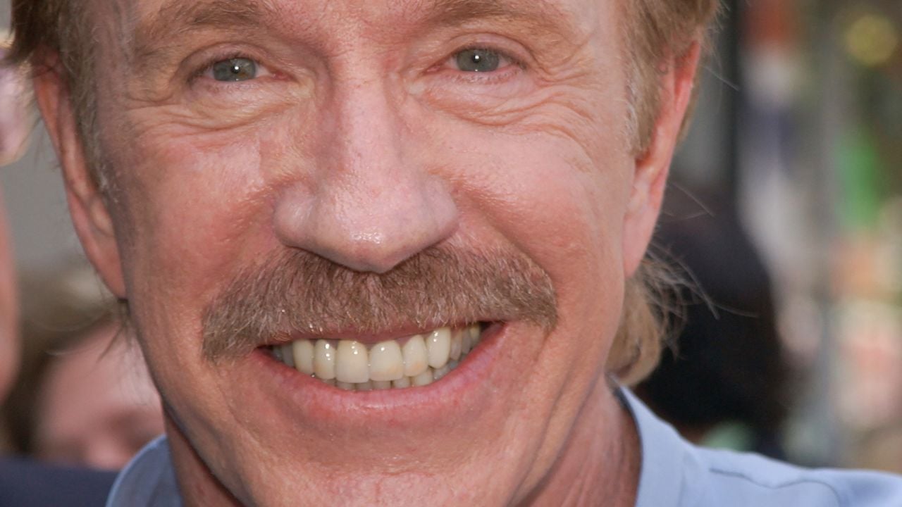A close-up of a smiling man with light skin, blue eyes, a mustache, and light brown hair. He is wearing a light blue collared shirt, and the background is blurred with people and outdoor light.