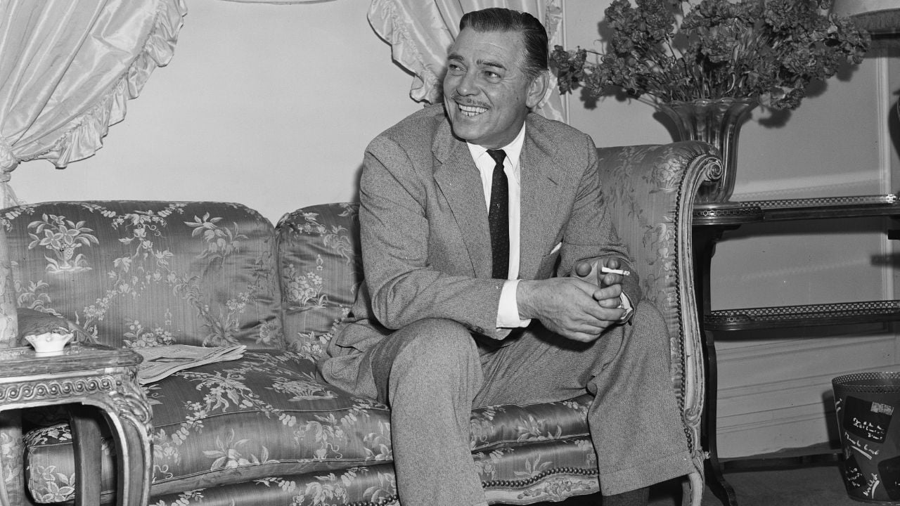 A man in a suit sits on a patterned sofa, smiling and holding a cigarette. There is a small table on the left and a vase of flowers on a table in the background. The setting appears to be a well-furnished room.