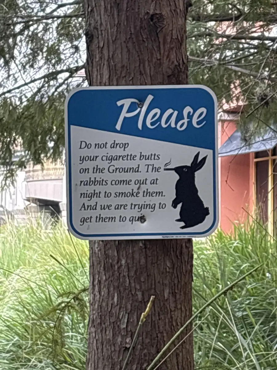 A humorous sign on a tree shows a rabbit silhouette and reads: “Please do not drop your cigarette butts on the ground. The rabbits come out at night to smoke them. And we are trying to get them to quit.”