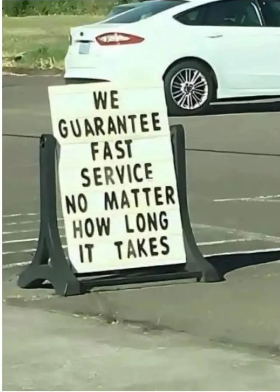 A sign in a parking lot reads, “We guarantee fast service no matter how long it takes,” with a white car parked in the background.