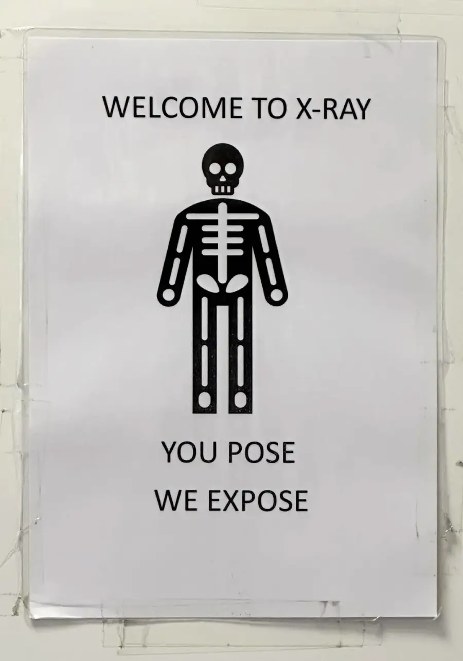 Sign with a cartoon skeleton, text above reads “WELCOME TO X-RAY,” below reads “YOU POSE WE EXPOSE.” The image humorously refers to X-ray imaging exposing the inside of the body.