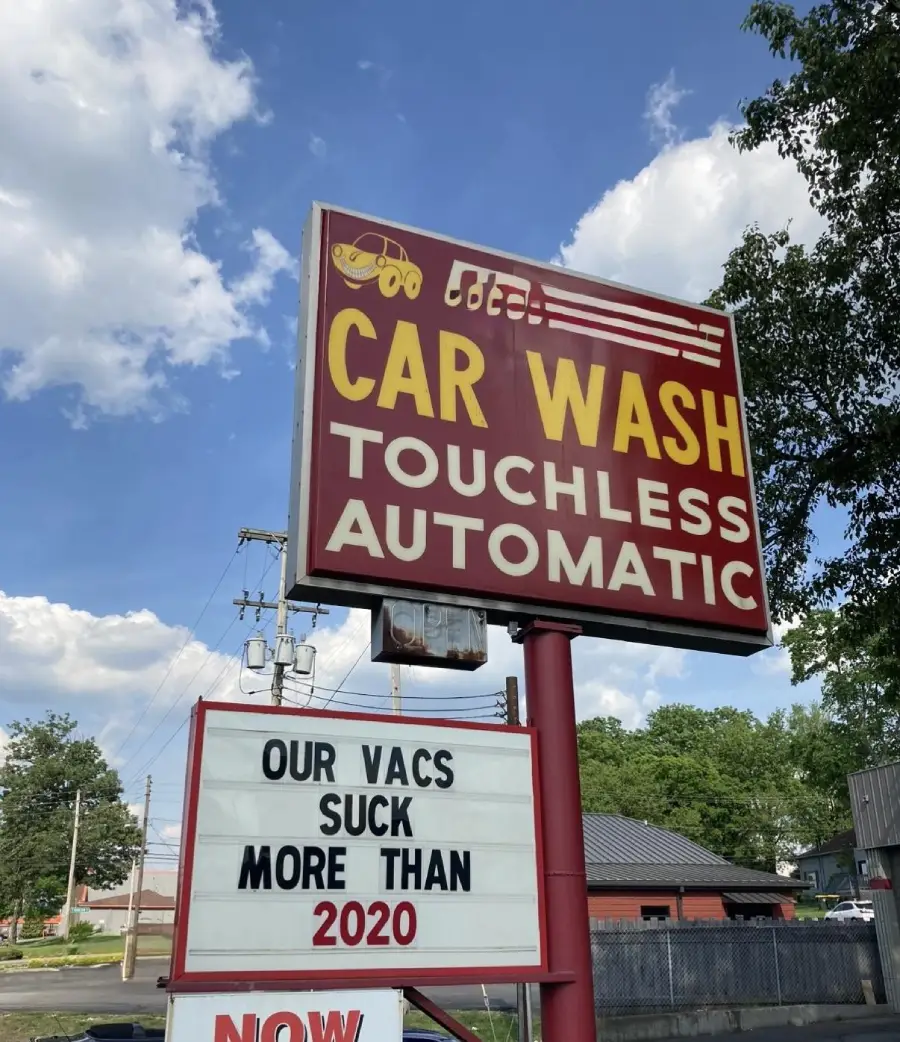 A car wash sign reads “OUR VACS SUCK MORE THAN 2020” under “CAR WASH TOUCHLESS AUTOMATIC” with a blue sky and trees in the background.