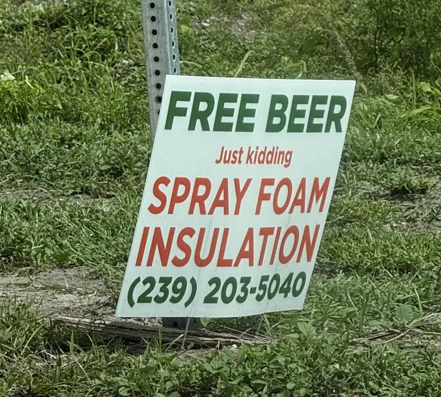 A sign on grass reads: “FREE BEER Just kidding SPRAY FOAM INSULATION (239) 203-5040.”