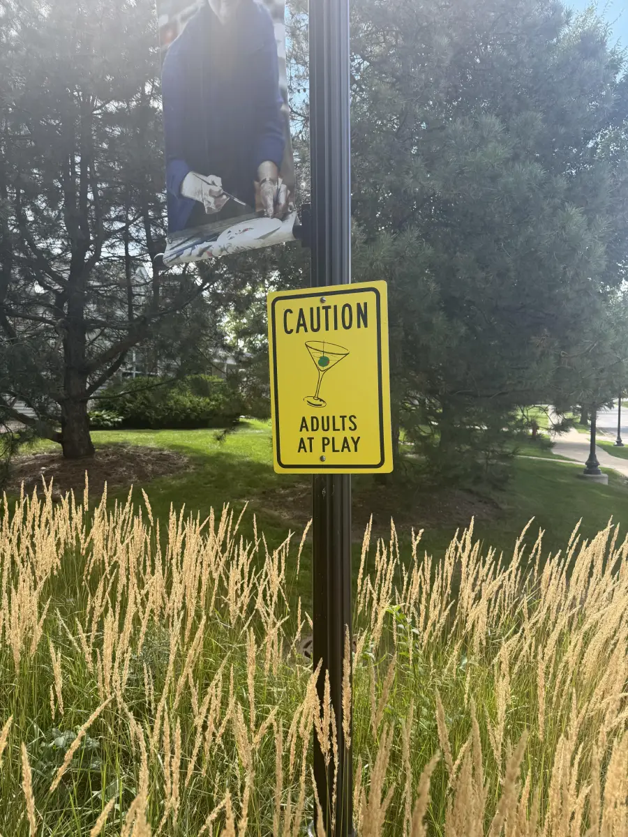 A yellow sign on a pole reads “Caution: Adults at Play” with a martini glass icon. Tall grass and trees are in the background.