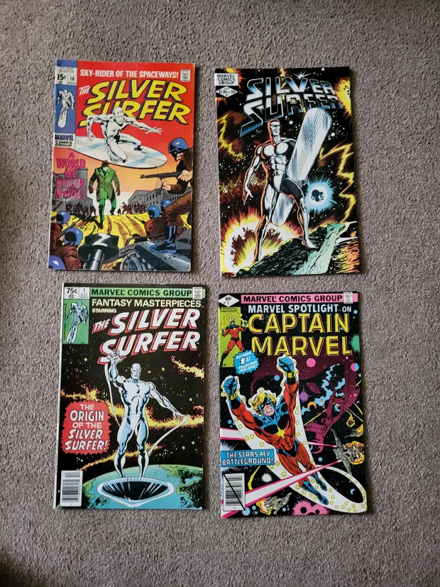 Four vintage Marvel comic books on carpet: three Silver Surfer issues featuring the character in various poses, and one Captain Marvel comic showing the hero with energy blasts and cosmic background.