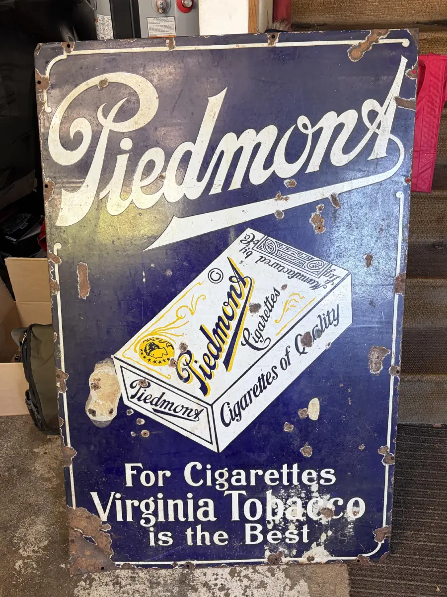 Vintage, weathered sign reading “Piedmont Cigarettes. For Cigarettes Virginia Tobacco is the Best.” Includes an illustration of a Piedmont cigarette box with visible wear and rust spots.