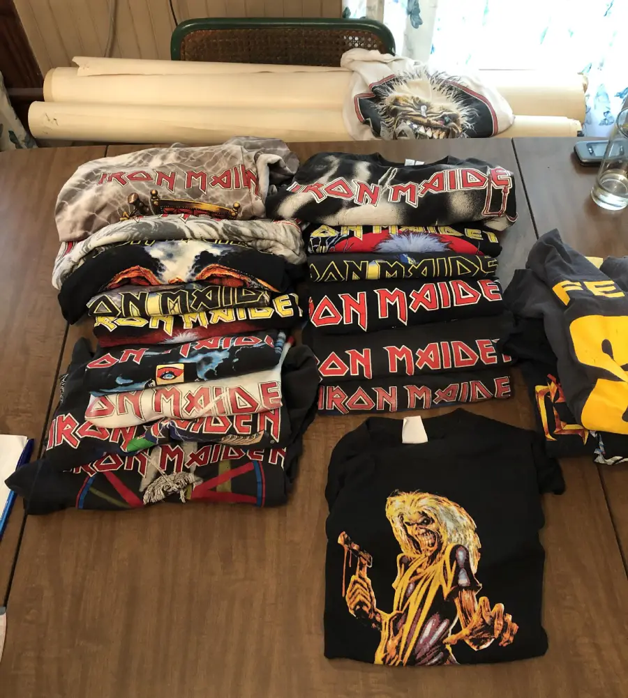 Nine folded Iron Maiden t-shirts with colorful logos and designs are arranged on a wooden table, along with one yellow and black shirt featuring a cartoon figure, and rolled white posters in the background.