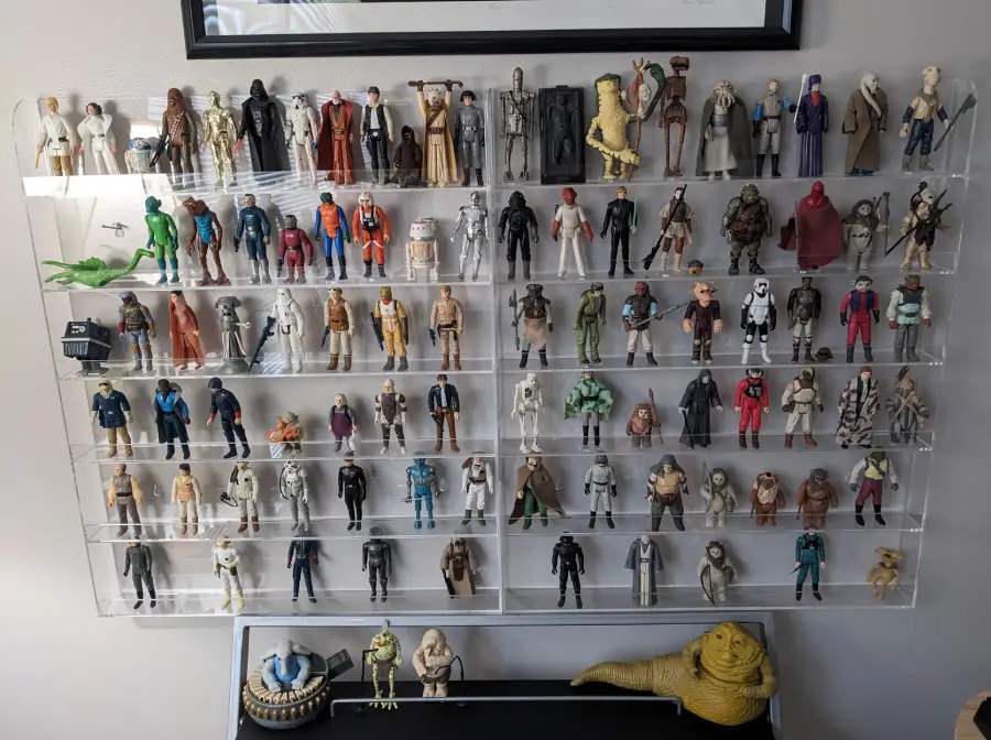 A display case with multiple shelves holding vintage Star Wars action figures, arranged in rows. A yellow Jabba the Hutt figure and accessories sit below the case on a black surface against a light wall.