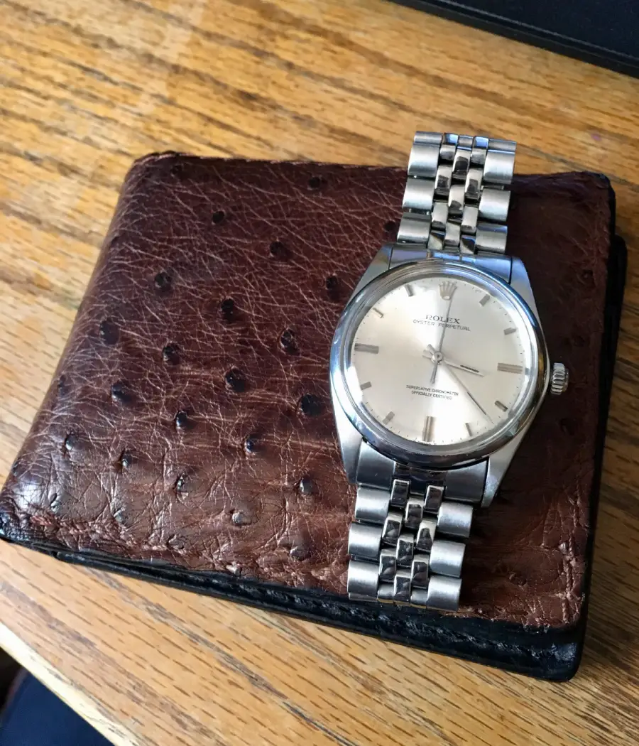 A silver wristwatch with a metal band rests on a brown, textured leather wallet with a dimpled pattern, placed on a wooden surface.