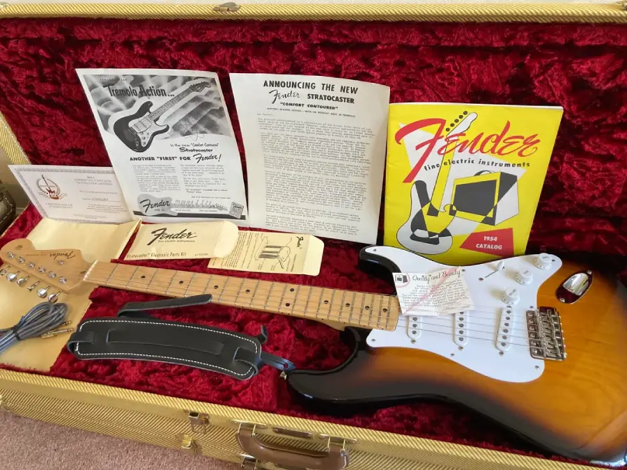 A sunburst Fender Stratocaster guitar in a red velvet-lined case, surrounded by vintage Fender booklets, care tags, manual, cable, and a black strap.