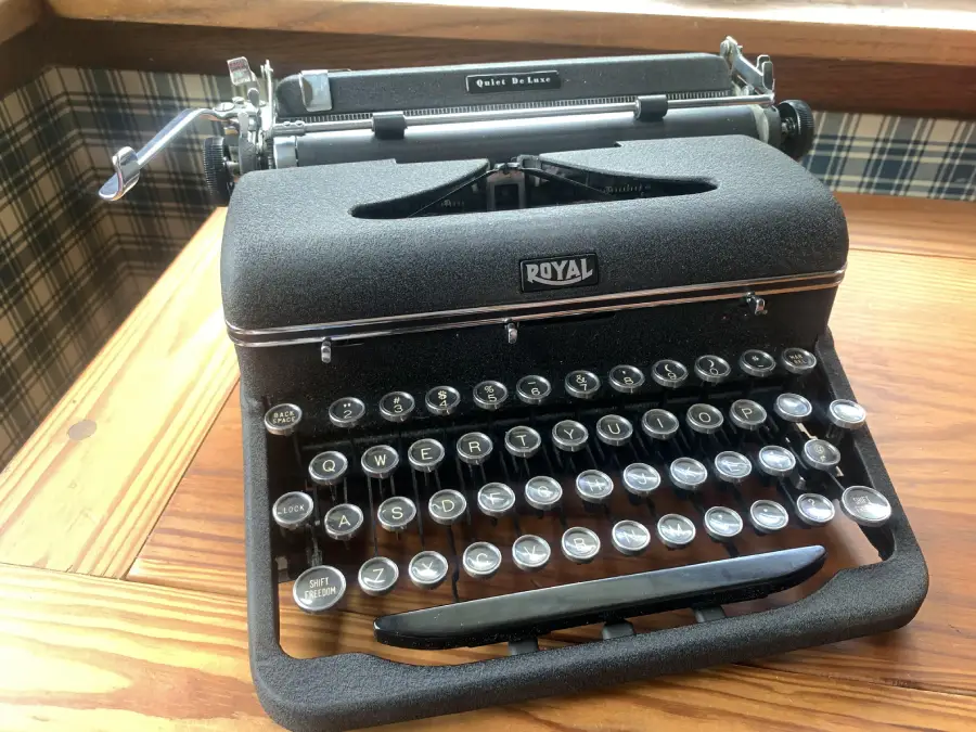 A vintage black Royal typewriter sits on a wooden desk by a window, with plaid wallpaper in the background.