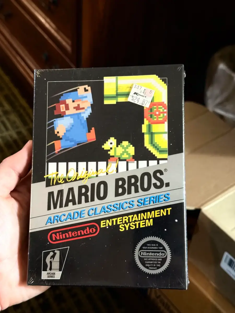 A hand holds a boxed copy of “Mario Bros. Arcade Classics Series” for the Nintendo Entertainment System, featuring retro artwork and price stickers on the plastic wrap.