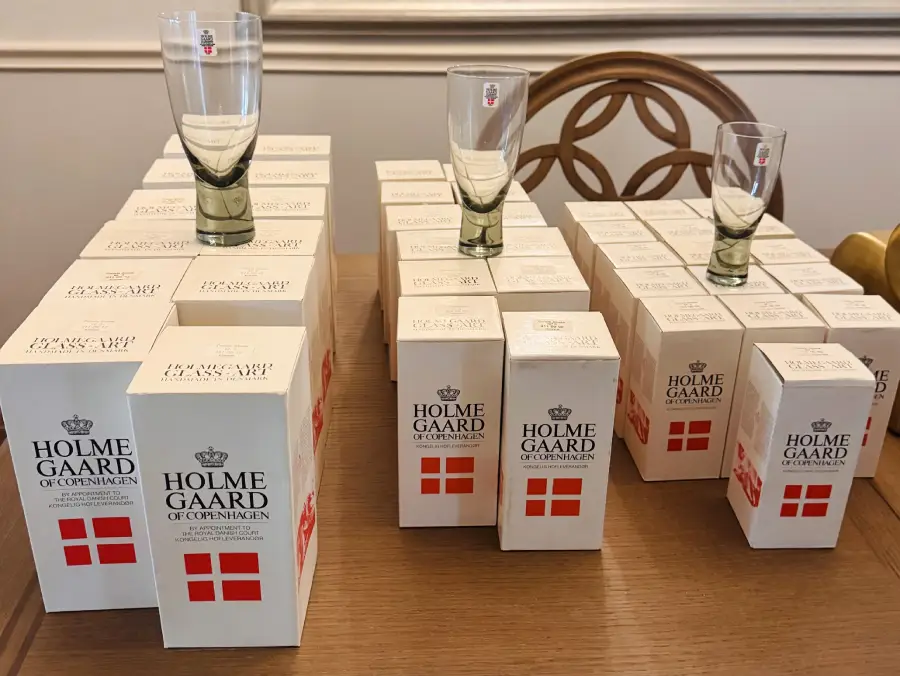 Several Holmegaard of Copenhagen glass boxes are arranged in rows on a table, with four empty drinking glasses placed on top of some of the boxes.