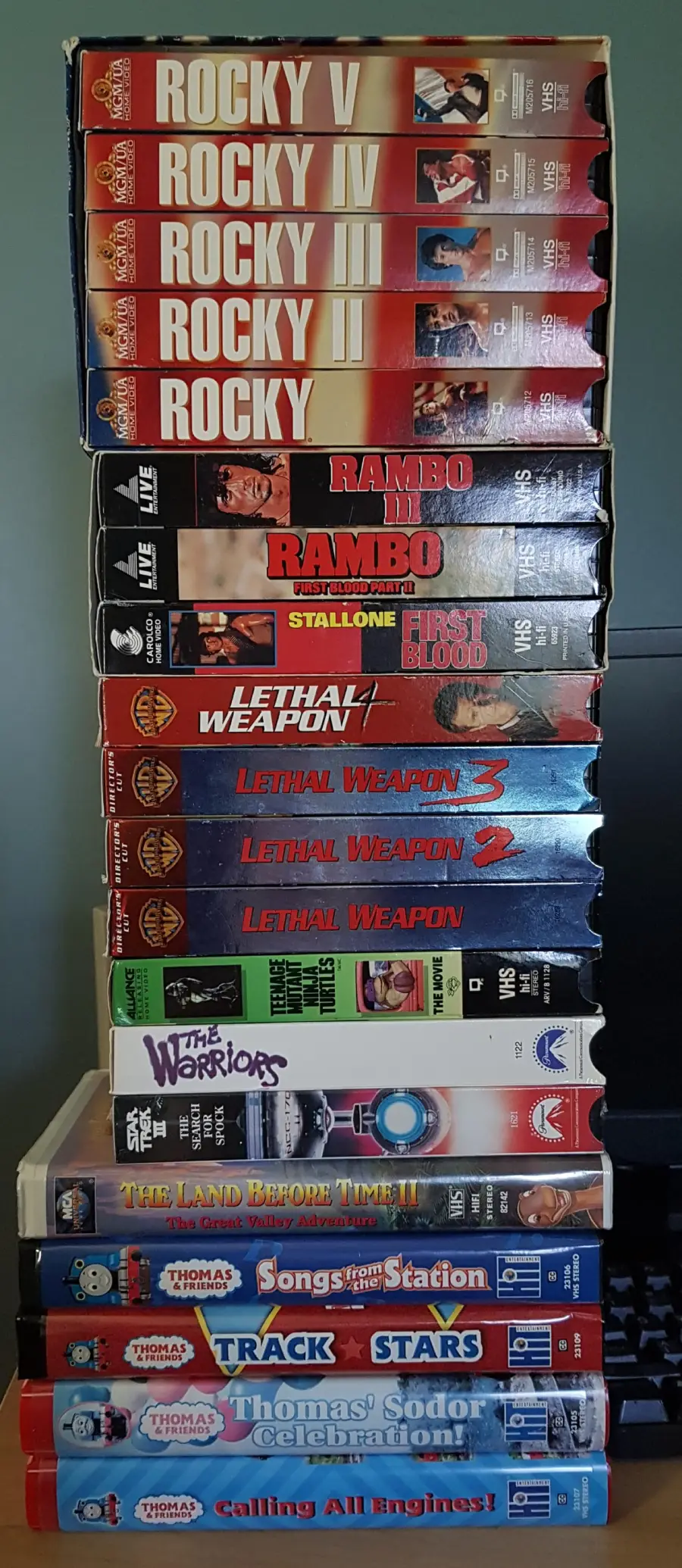 Stack of VHS tapes, including Rocky films, Rambo, Lethal Weapon series, The Warriors, Robocop, The Land Before Time, Thomas & Friends, and other children’s movies, arranged on a shelf against a plain background.