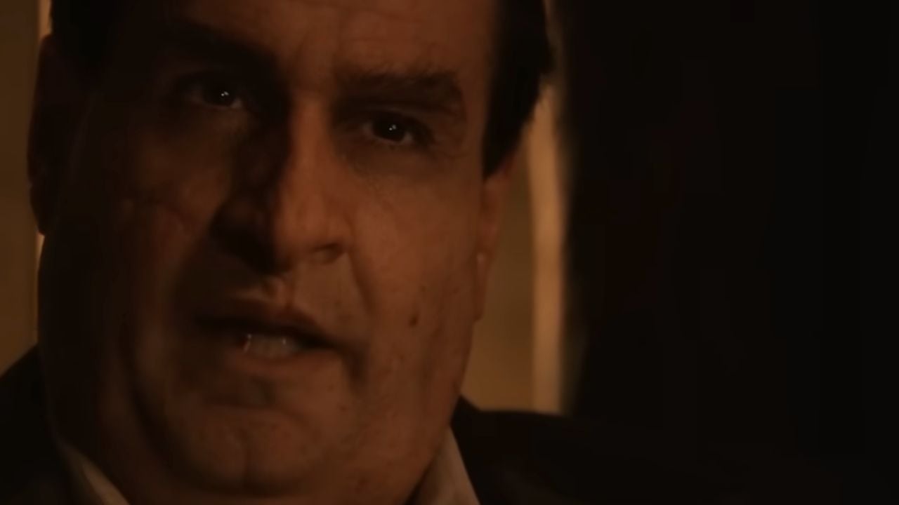 A close-up of a man with a serious expression, partially in shadow, lit by warm, dim lighting.