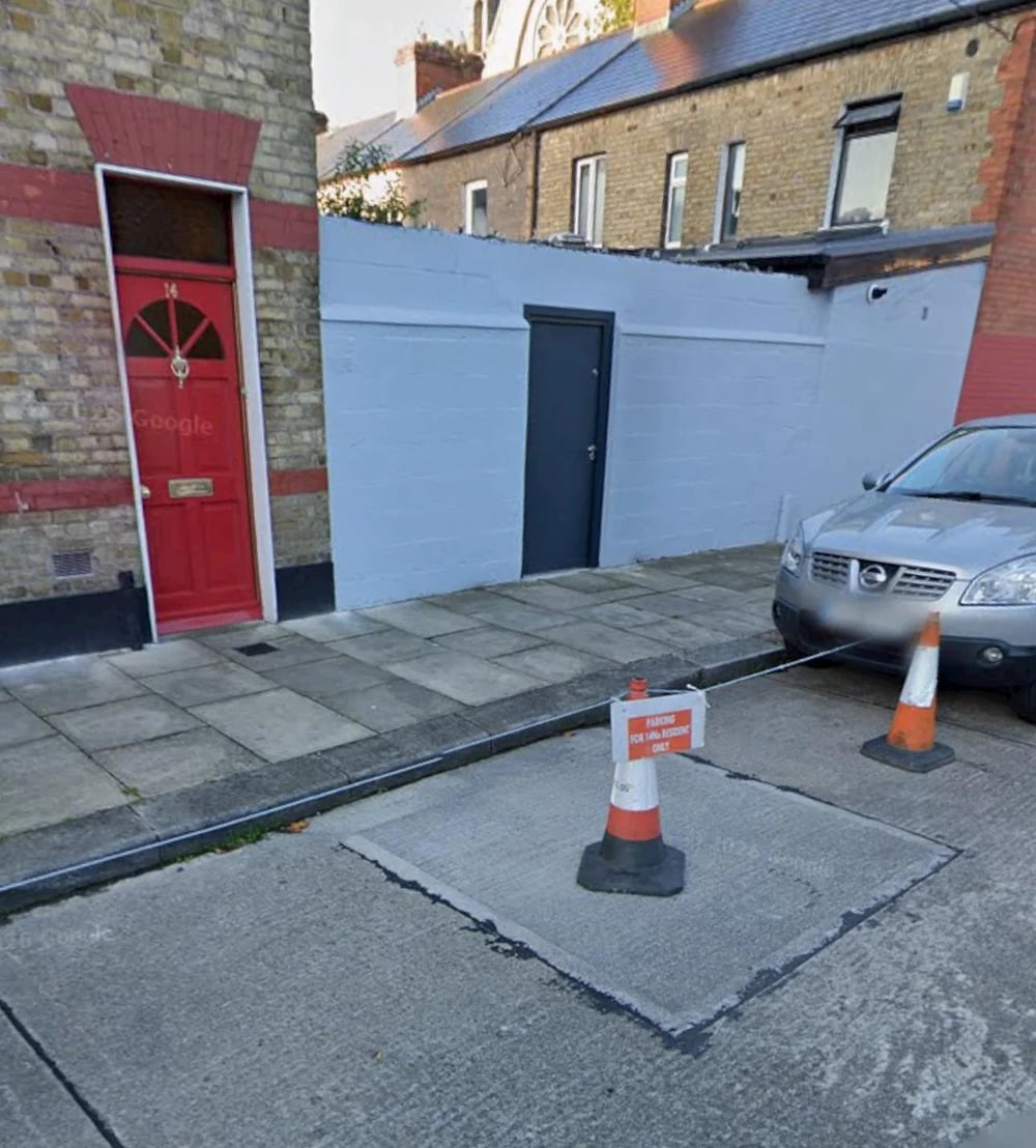 A sidewalk and street with a red door, a gray door, a parked car, and traffic cones around a metal cover labeled “NO PARKING 24hr Access Required” in front of the house.