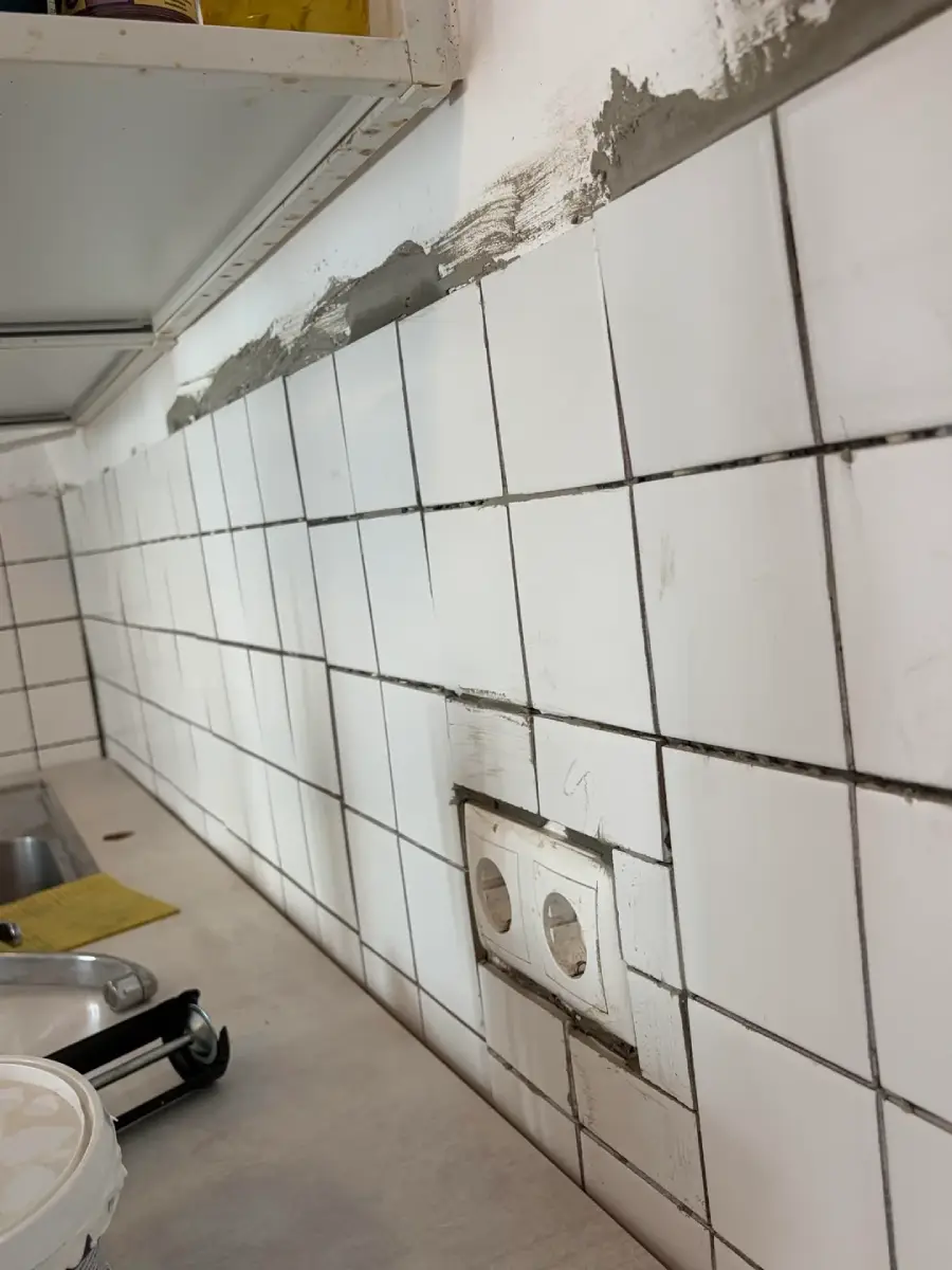 White kitchen wall tiles being installed, with some tiles missing grout and some wall areas unfinished. A double electrical outlet is visible among the tiles above the counter.