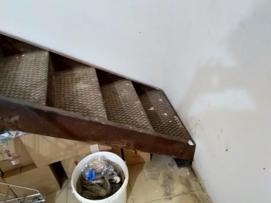 A metal staircase ends abruptly at a wall, with boxes and a white bucket on the floor below. The stairway appears unusable as it leads directly into the wall.