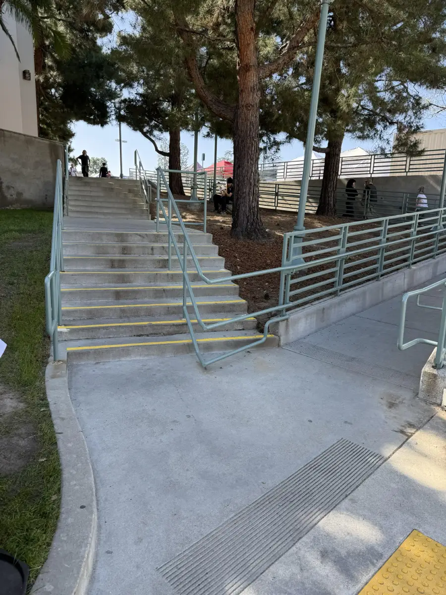 A concrete staircase with yellow edges and a ramp with railings run parallel under tall pine trees; a tactile paving section is in the foreground.