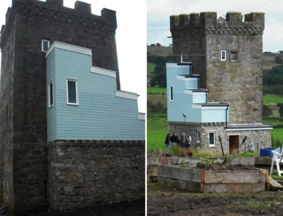 A medieval stone tower with a modern, light blue wooden extension attached to one side, set in a rural landscape with hills and greenery in the background.