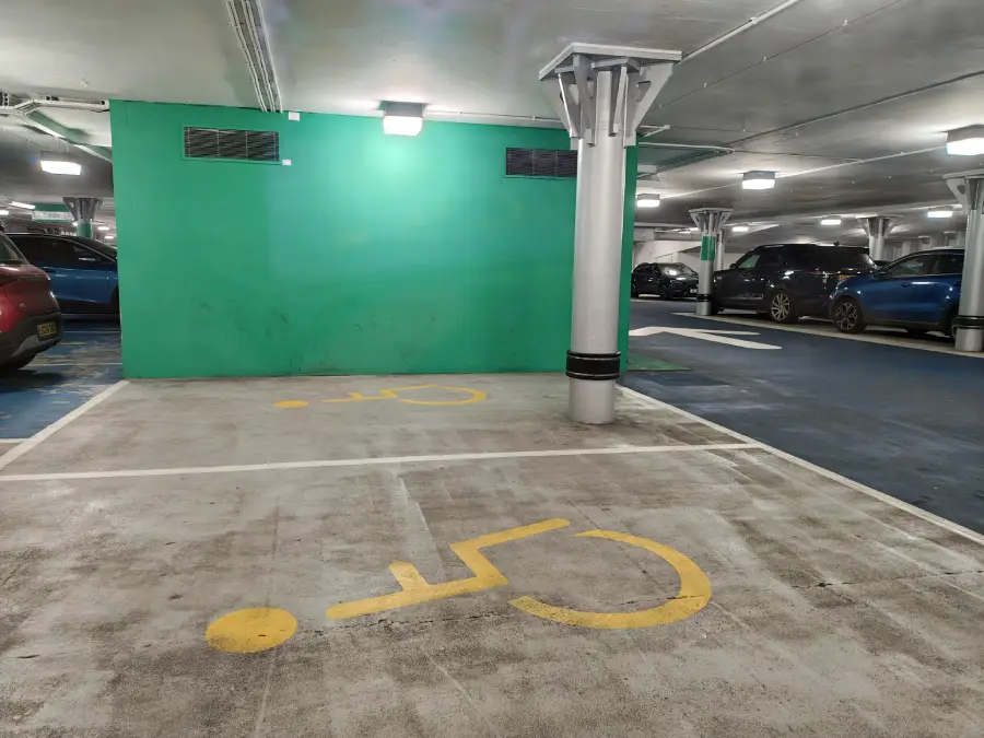 Two accessible parking spaces in an indoor lot are blocked by a large column, making them unusable. The area has yellow wheelchair symbols painted on the ground and a green wall in the background.