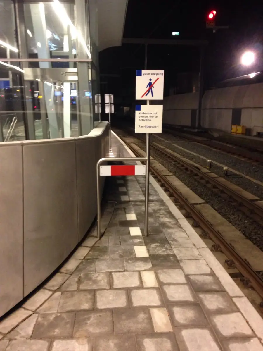 A narrow pedestrian path at a train station is blocked by a metal barrier and a sign that indicates no entry. Train tracks run alongside the path.