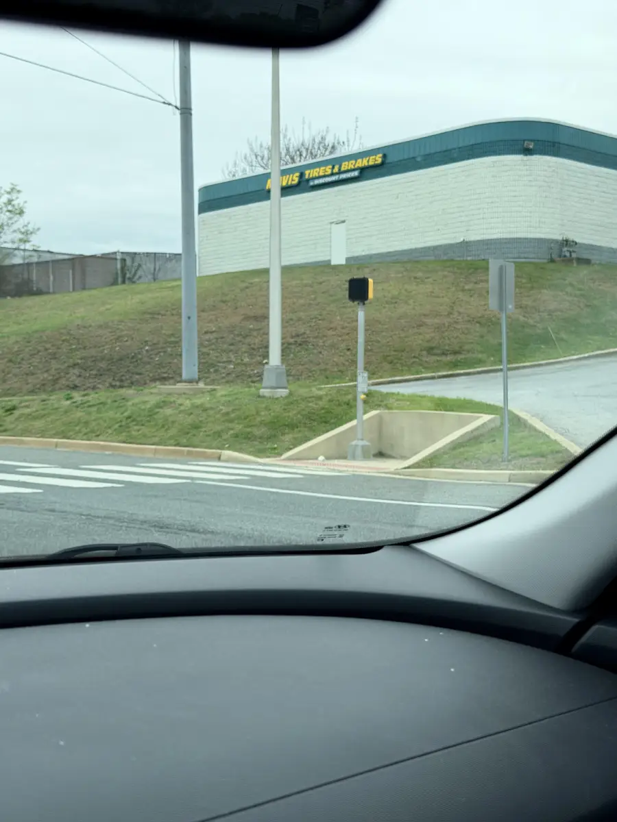 View from a car shows a sloped grassy area with a sidewalk leading directly into a curb, ending abruptly. A building with a “TIRES & BRAKES” sign is in the background.
