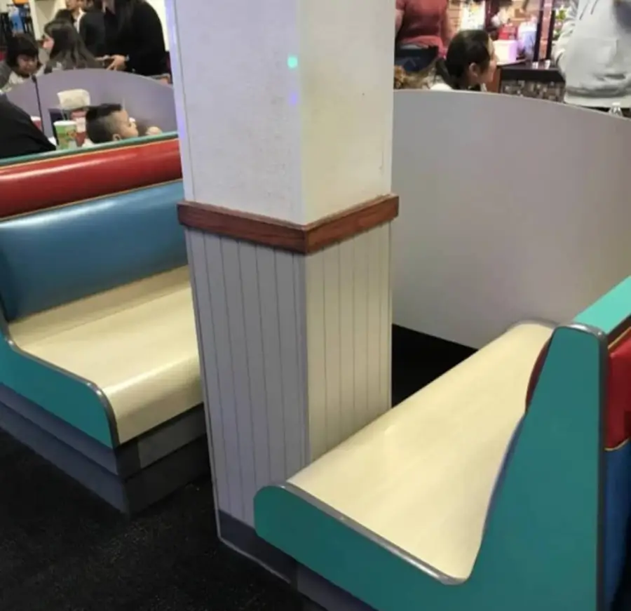 Two restaurant booths are divided by a wall, making it impossible to sit at either table. People and children are visible in the background.