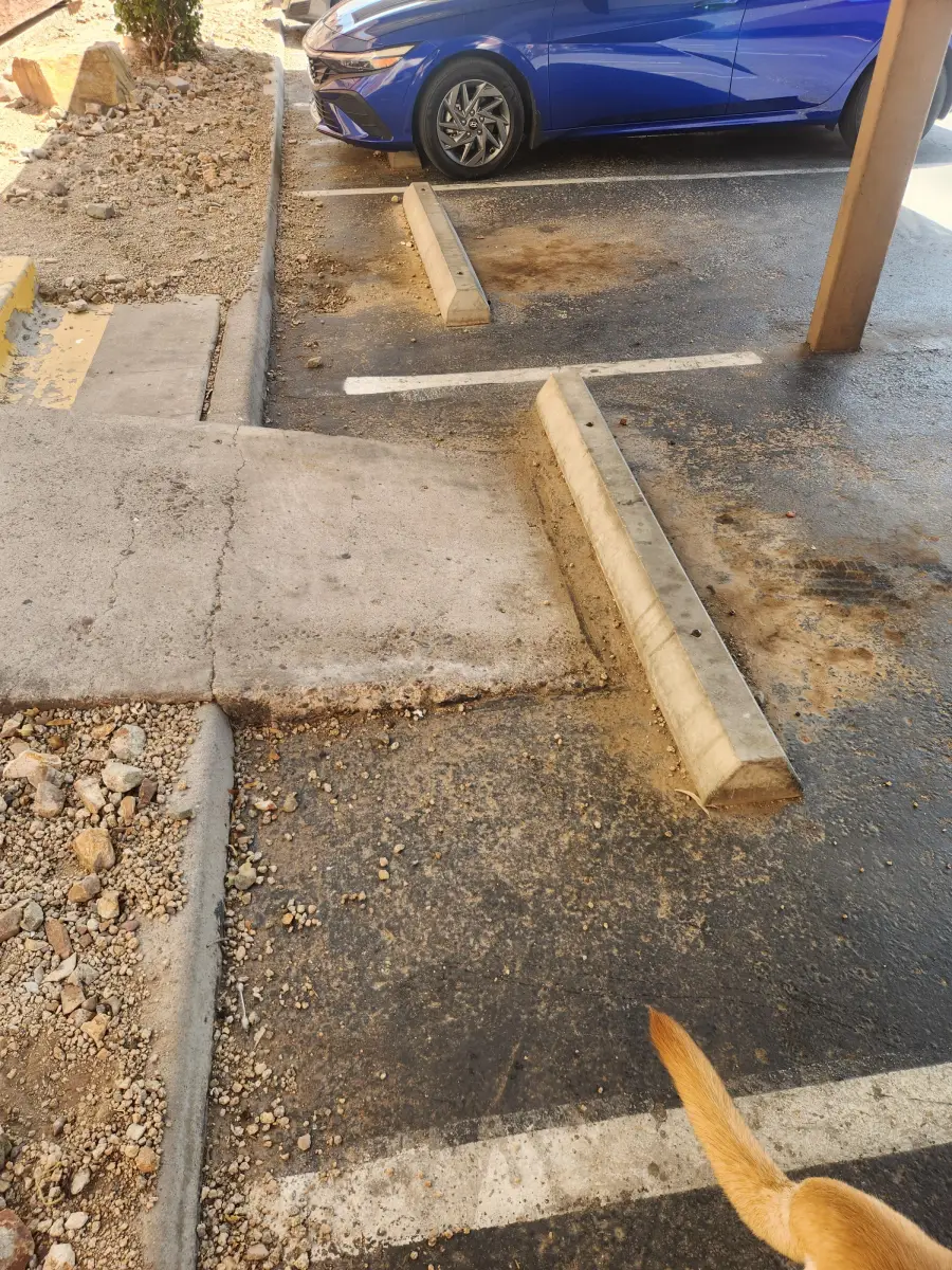 A blue car is parked in a parking spot next to a concrete ramp and curb. Gravel and dirt cover parts of the ground. The tip of a dog’s tail is visible at the lower right corner.