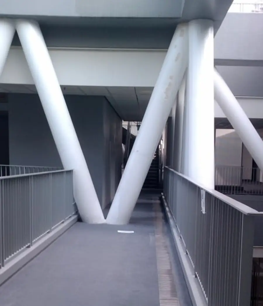 A walkway is blocked by large white diagonal beams, preventing passage. Gray metal railings line the sides, and stairs are visible in the background.