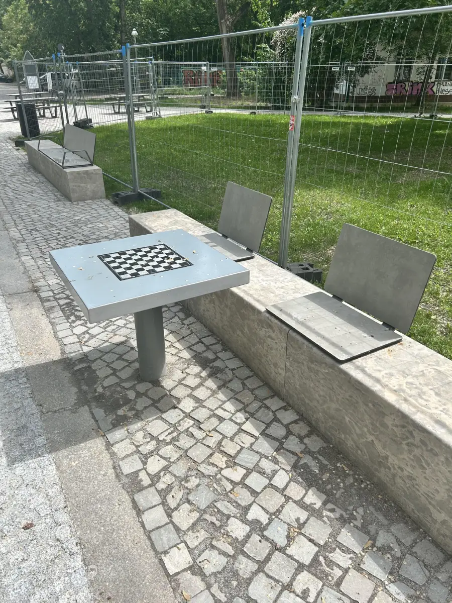 Outdoor chess table with built-in metal seats, beside a stone bench on a cobblestone path. Metal fencing and grass are in the background, with sunlight casting shadows.