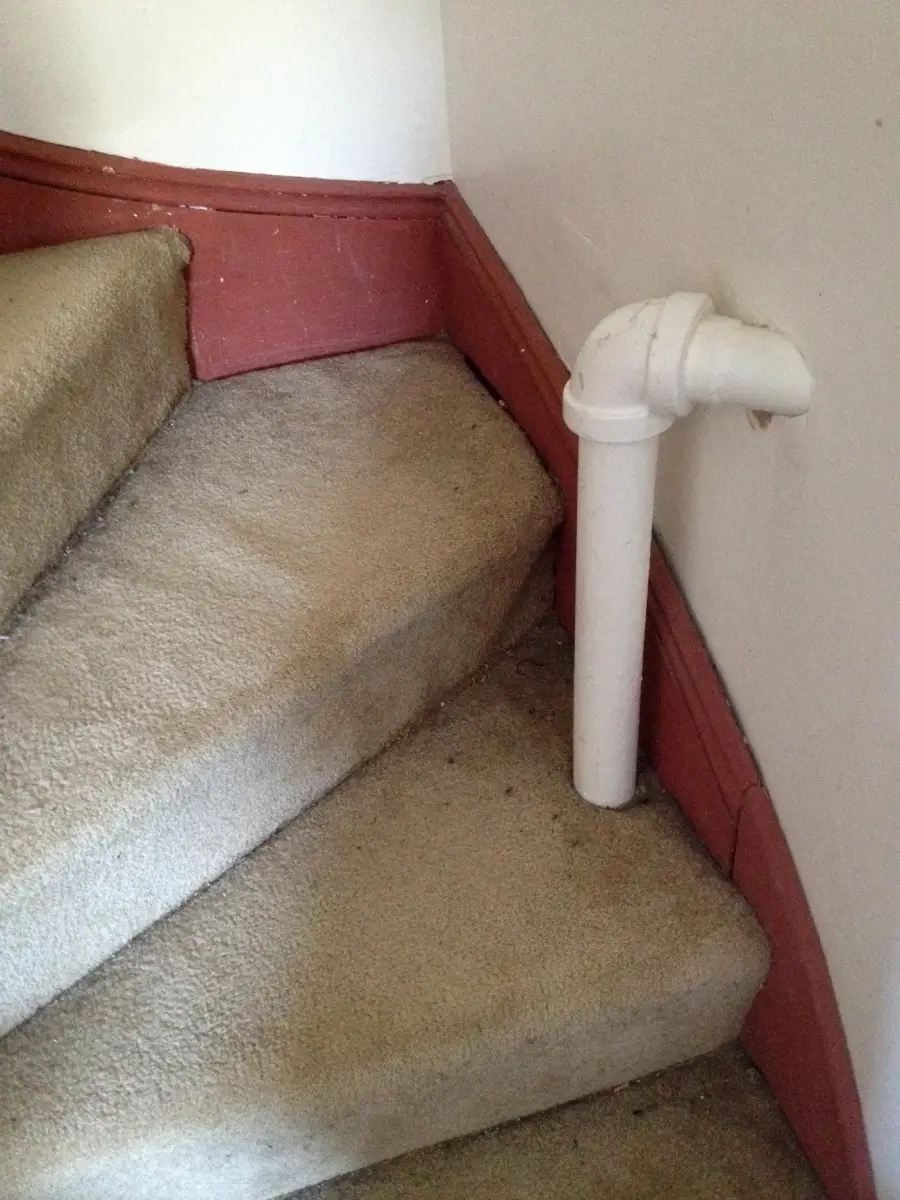 A carpeted staircase with a white PVC pipe protruding vertically from one step near the wall, obstructing part of the pathway up the stairs.
